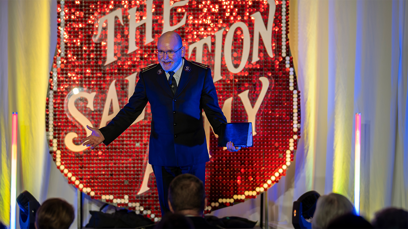 General Lyndon Buckingham, holding a Bible, speaks to the crowd in front of a large Salvation Army red shield