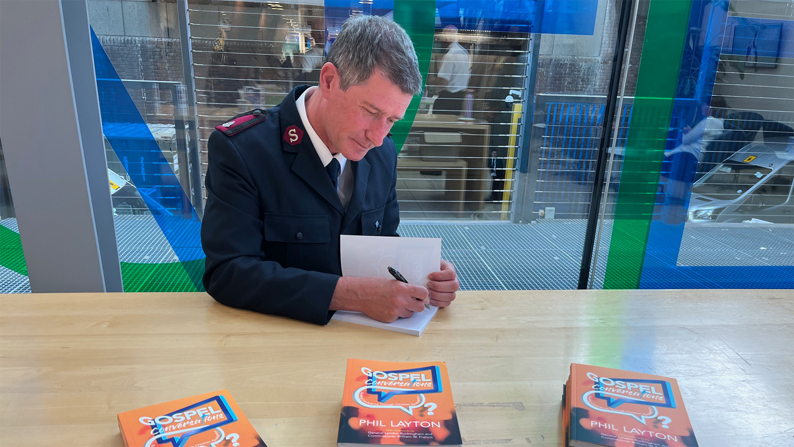Major Phil Layton, in Salvation Army uniform, sits at a wooden table signing a copy of his new book, Gospel Conversations