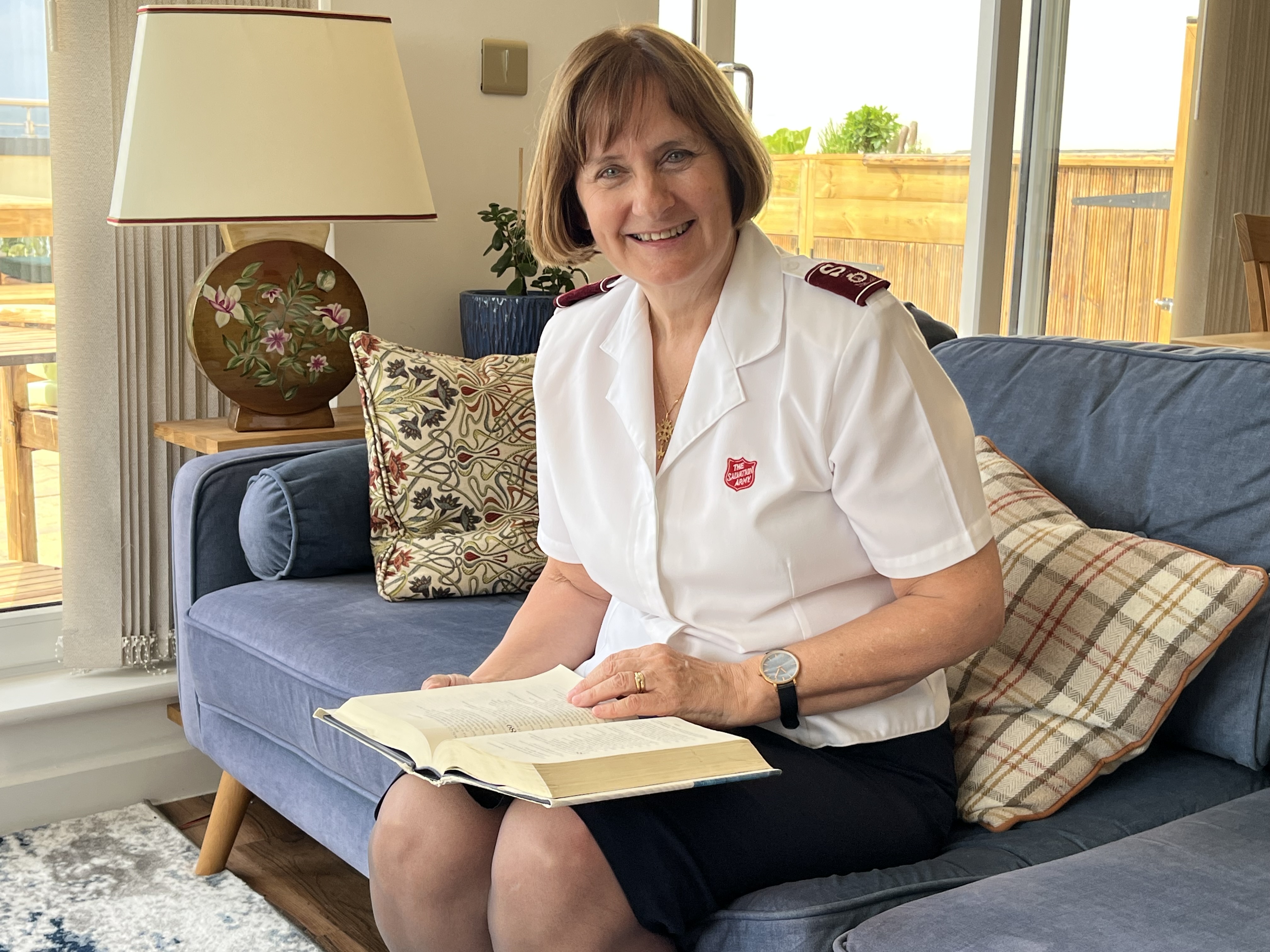 Commissioner Paone is sitting on her sofa holding a Bible, smiling at the camera