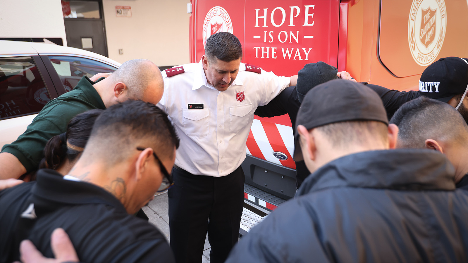Eight people including a Salvation Army officer stand in a circle, hands around each others' shoulders and heads bowed in prayer. In the background a Salvation Army truck shows the message 'Hope is on the way'.