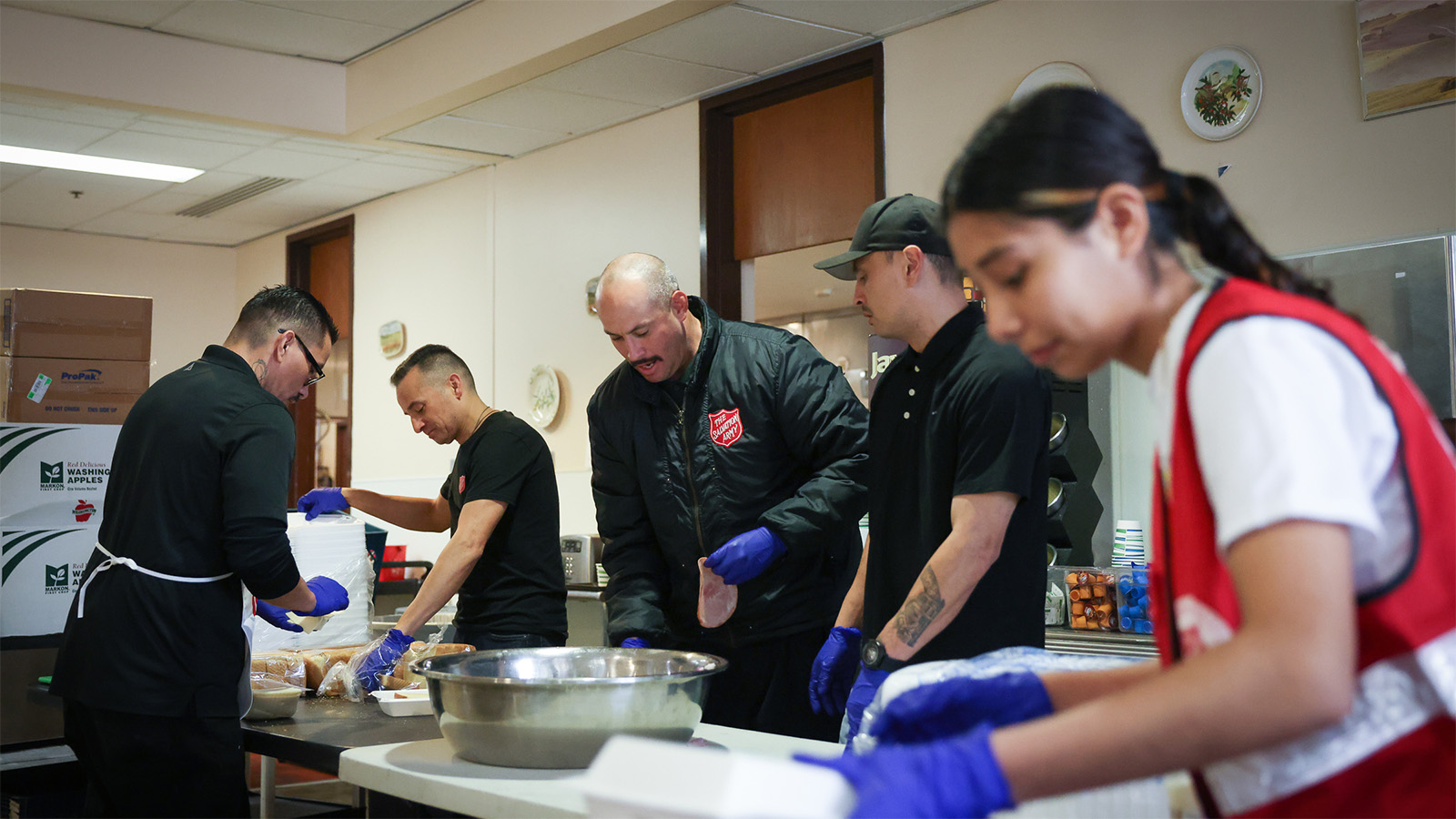 A group of people wearing clothes with the Salvation Army Red Shield prepare sandwiches and pack them into takeaway boxes