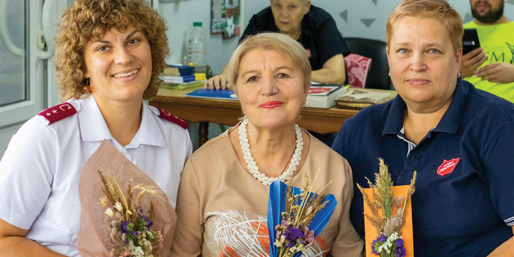 Three women, two wearing Salvation Army clothing, hold flower bouquets