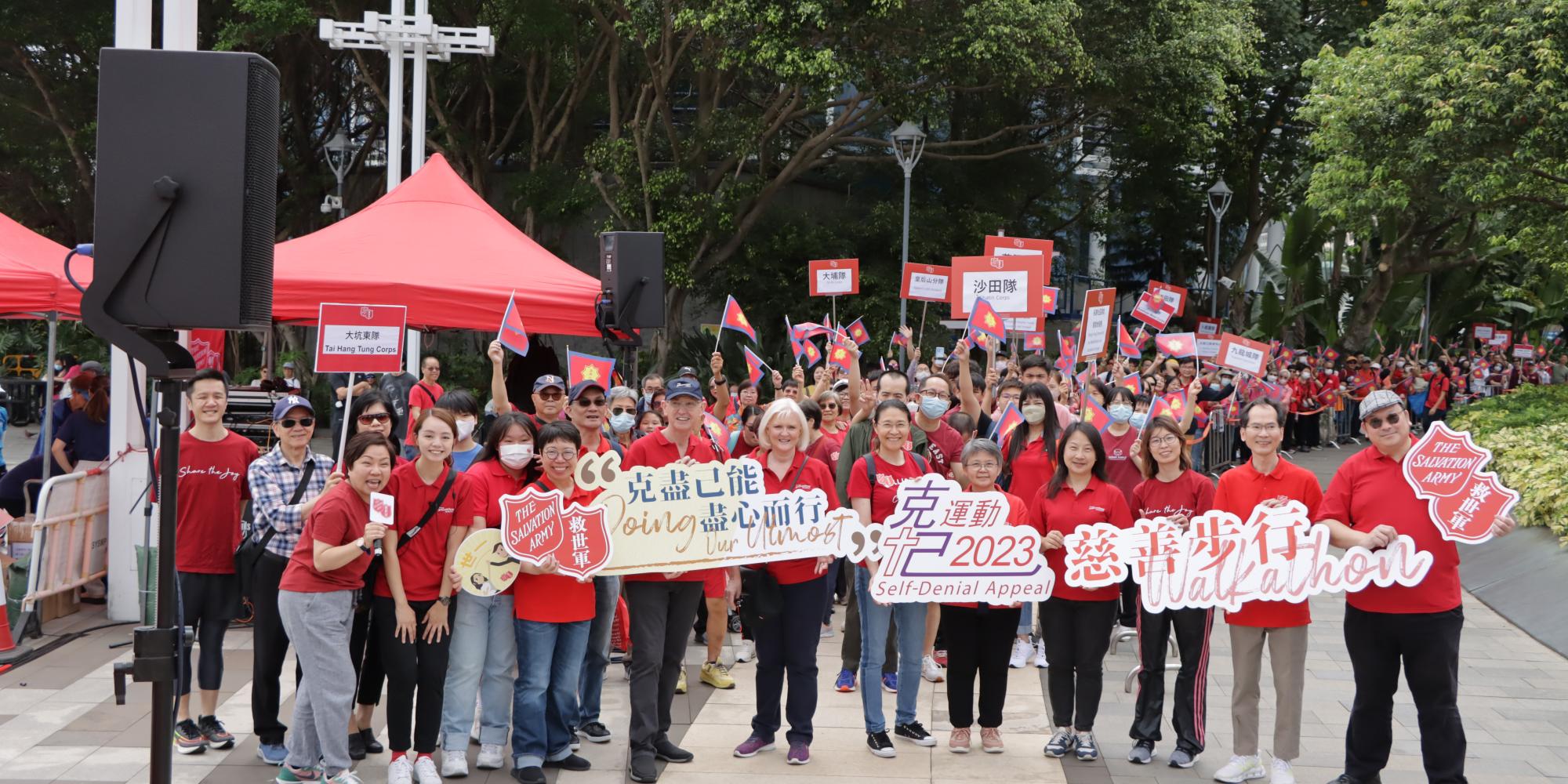 A large group of people in red t-shirts hold banners with the Red Shield and message '2023 Self-Denial Appeal'