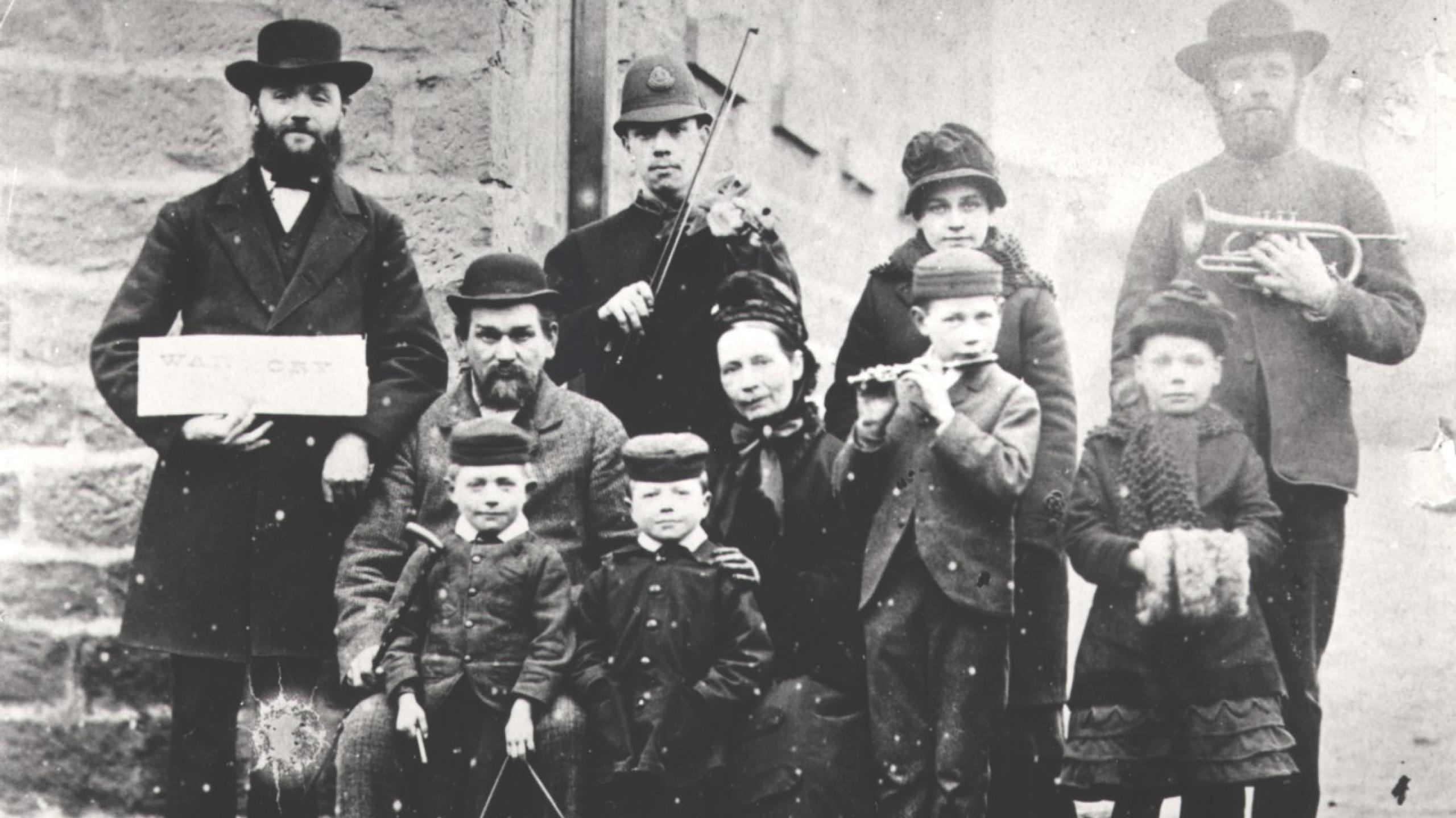 A group of adults and children, one with a violin, one with a cornet, one with a triangle and one with a flute