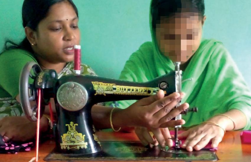 Two women sitting at a sewing machine, one with her face blurred