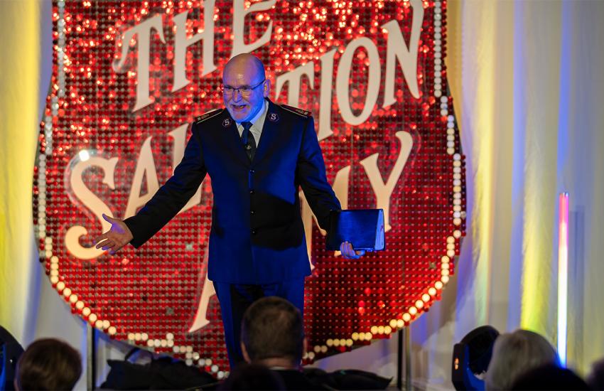 The General addresses a crowd in front of a large Red Shield