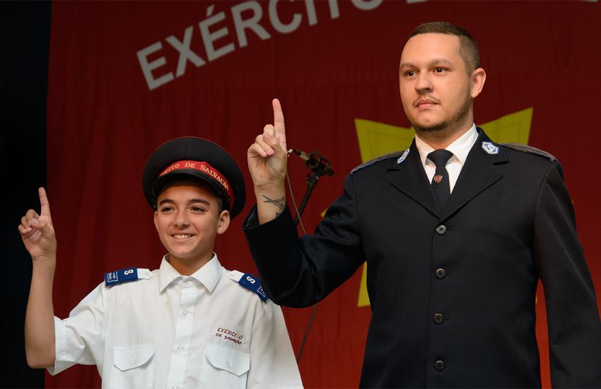 A young junior soldier, wearing a cap, and a senior soldier give the Salvation Army salute