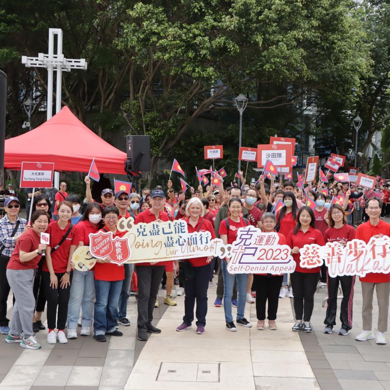 A large group of people in red t-shirts hold banners with the Red Shield and message '2023 Self-Denial Appeal'