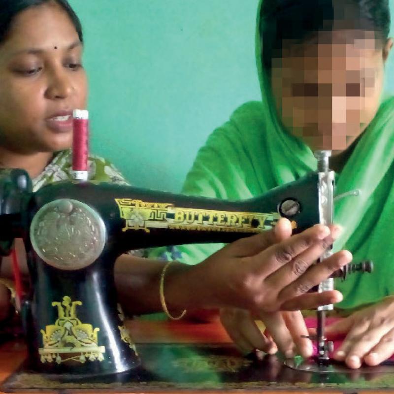 Two women sitting at a sewing machine, one with her face blurred