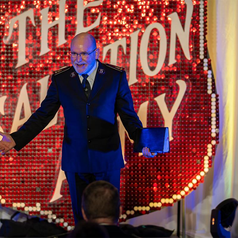 The General addresses a crowd in front of a large Red Shield